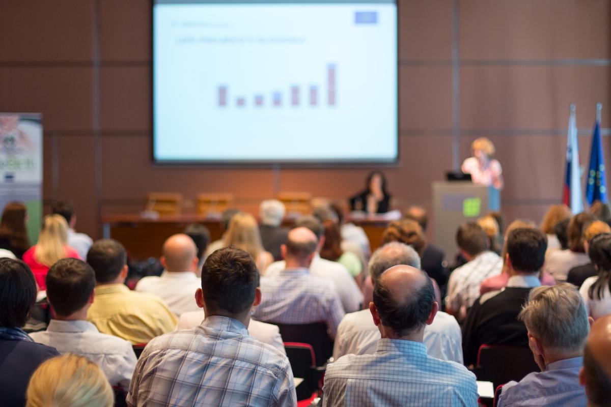 A speaker gives a talk at a conference to a crowd of people.